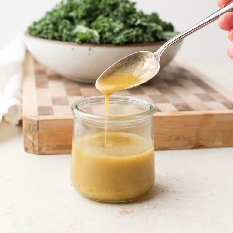 A jar of homemade vinaigrette with vinaigrette dropping off a spoon into the jar in front of a cutting board with a bowl of kale.
