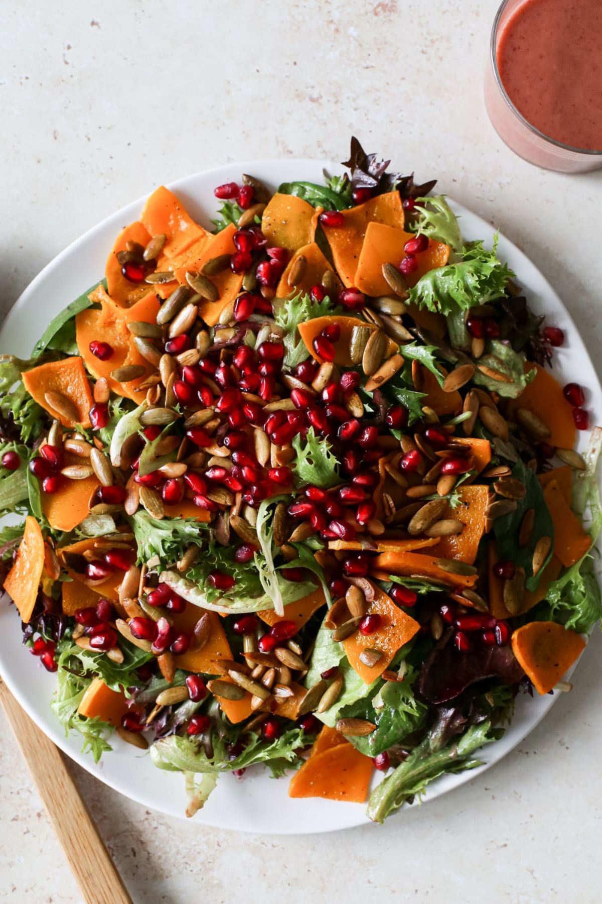 A large plate of fall harvest salad with mixed spring greens, roasted butternut squash slices, pomegranate seeds, and toasted pumpkin seeds next to a cranberry vinaigrette.