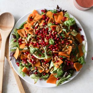 A large plate of fall harvest salad with mixed spring greens, roasted butternut squash slices, pomegranate seeds, and toasted pumpkin seeds next to a cranberry vinaigrette.