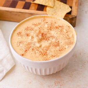 A bowl of cashew queso with green chiles next to a cutting board with tortilla chips.