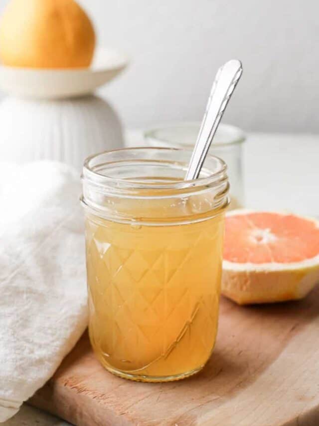 A jar with a metal spoon of grapefruit simple syrup on a brown wooden cutting board next to a half of pink grapefruit and a whole grapefruit.