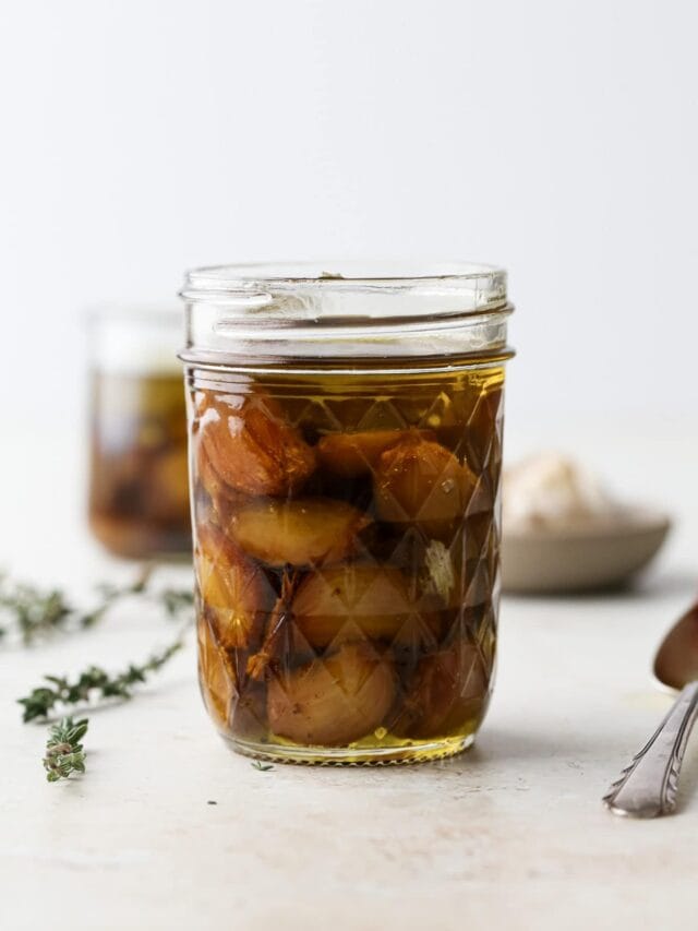 A glass jar of oven roasted garlic confit next to fresh thyme sprigs and a silver spoon.