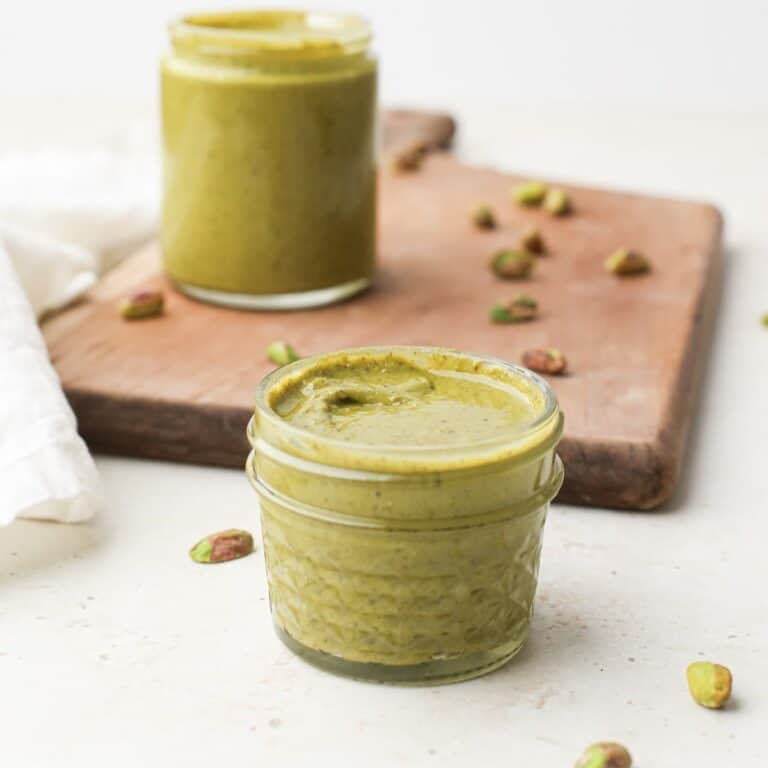Two jars of homemade pistachio butter on a cutting board with shelled pistachios.