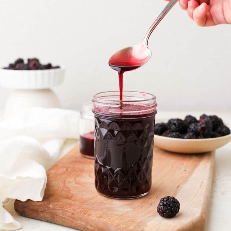 A spoonful of blackberry simple syrup dripping off a spoon into a jar next to fresh blackberries.