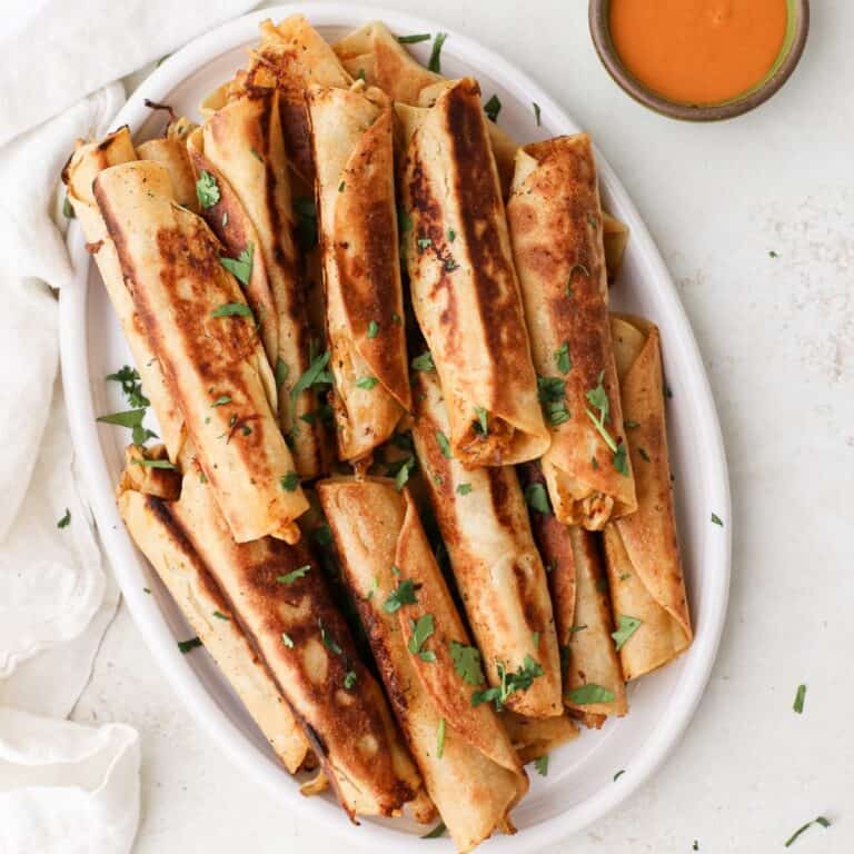 A plate of dairy free buffalo chicken taquitos topped with cilantro and served with a side of dairy free ranch and buffalo sauce.