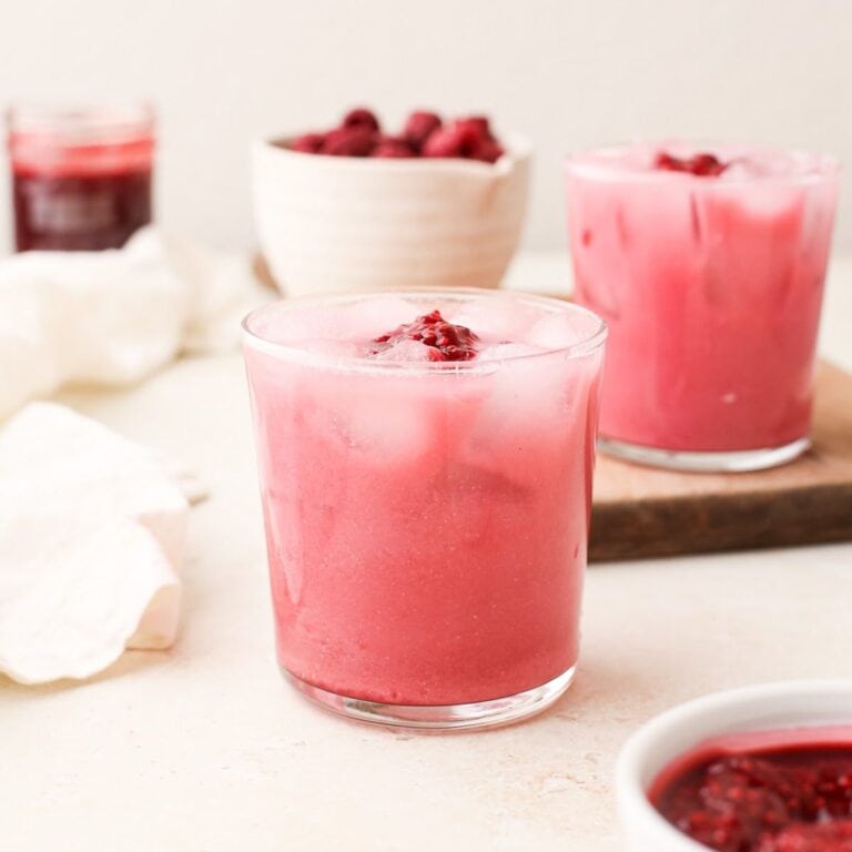 Two glasses of raspberry cream soda with raspberry puree on top next to a bowl of raspberries.