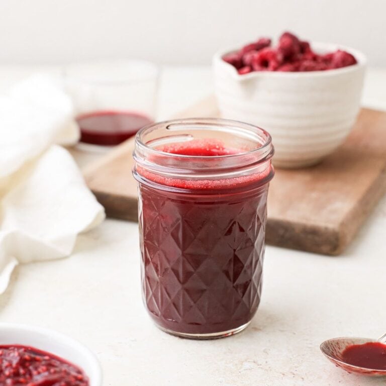A jar of homemade raspberry syrup next to a bowl of frozen raspberries.