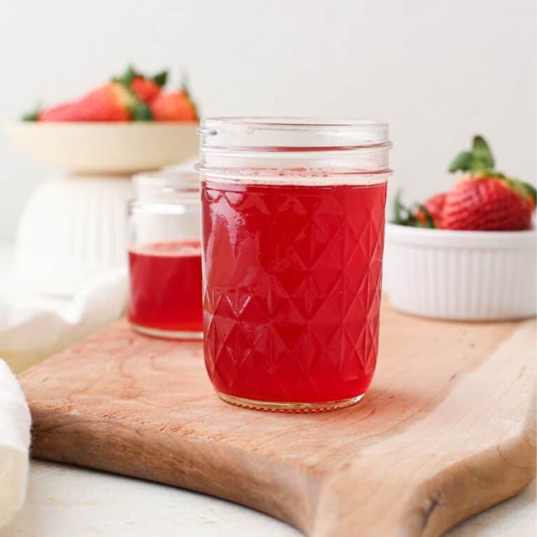 Two jars of homemade strawberry simple syrup next to two bowls of fresh strawberries.