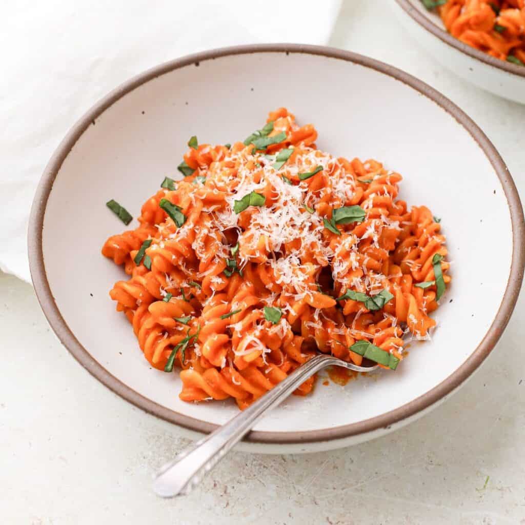 A bowl of dairy free creamy roasted red pepper pasta topped with grated dairy free parmesan cheese and torn basil leaves.