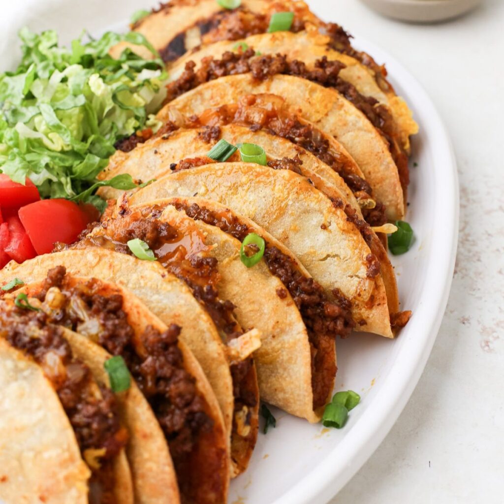 Crispy beef tacos overlapping each other on a serving plate with shredded lettuce and diced tomatoes.