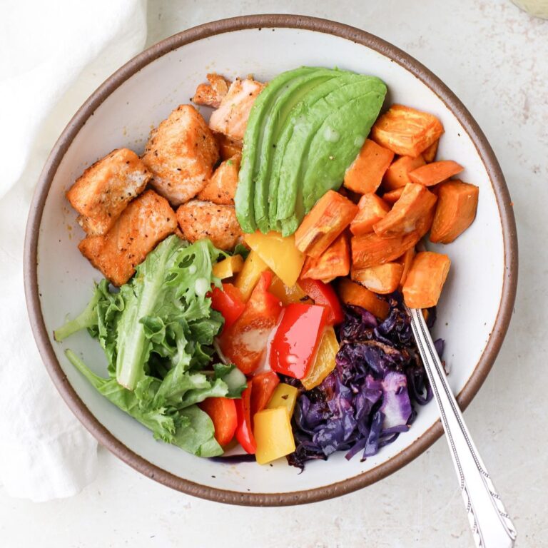 Salmon buddha bowl with roasted sweet potato, roasted purple cabbage, leafy greens, roasted colorful bell peppers, sliced avocado and cooked salmon cubes.