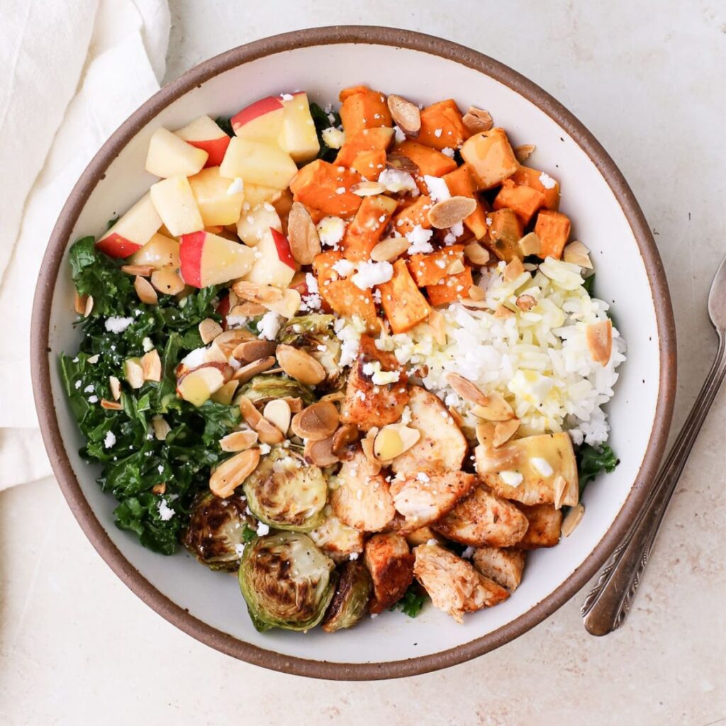 Sweetgreen inspired harvest bowl with sweet potatoes, massaged kale, chopped apples, chicken, rice, dairy free feta cheese, toasted almonds, and orange dressing.