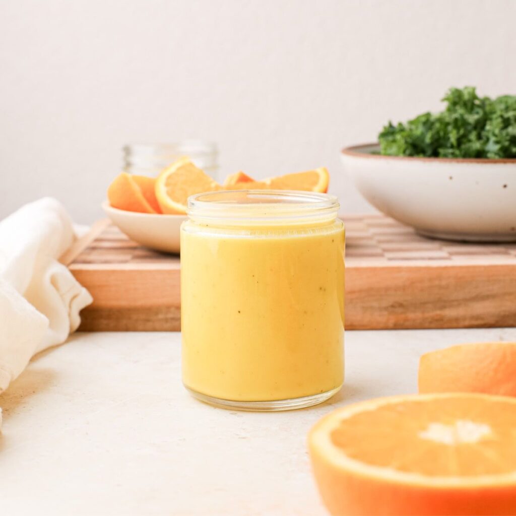 A jar of homemade orange vinaigrette next to fresh oranges and a bowl of massaged kale.