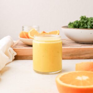 A jar of homemade orange vinaigrette next to fresh oranges and a bowl of massaged kale.
