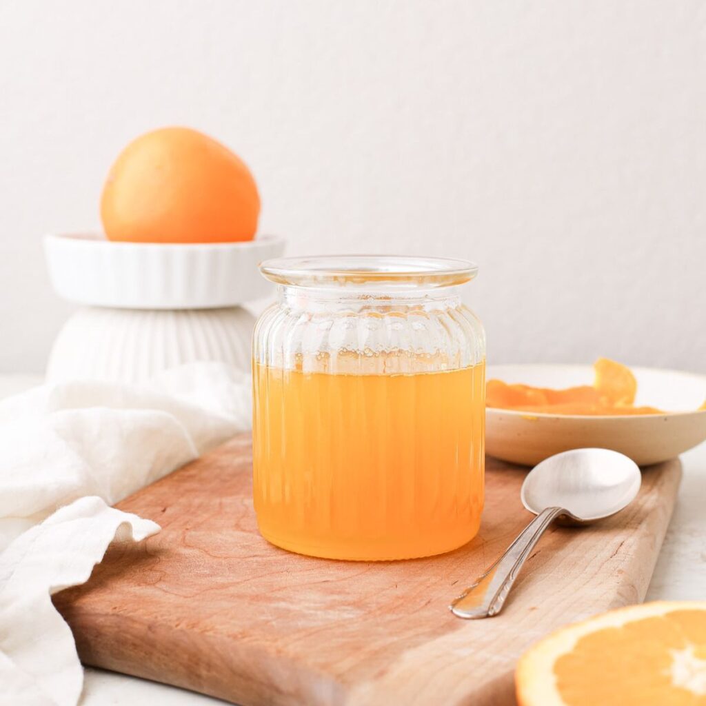 A jar of homemade orange simple syrup on a wooden cutting board next to fresh oranges.
