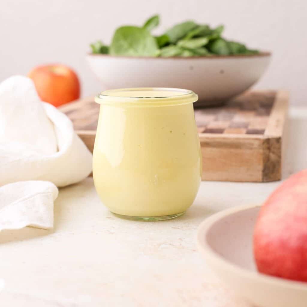A jar of homemade fresh apple vinaigrette dressing next to fresh apples and a bowl of greens.