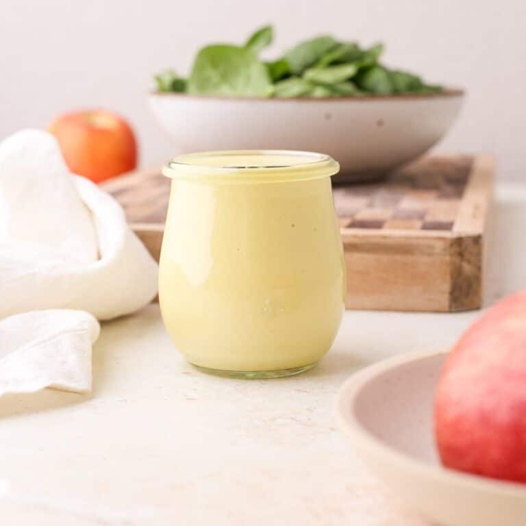 A jar of homemade fresh apple vinaigrette dressing next to fresh apples and a bowl of greens.