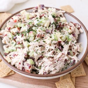 A bowl of cranberry pecan chicken salad with celery, apple, and a sprinkle of fresh dill on top next to crackers on a cutting board.