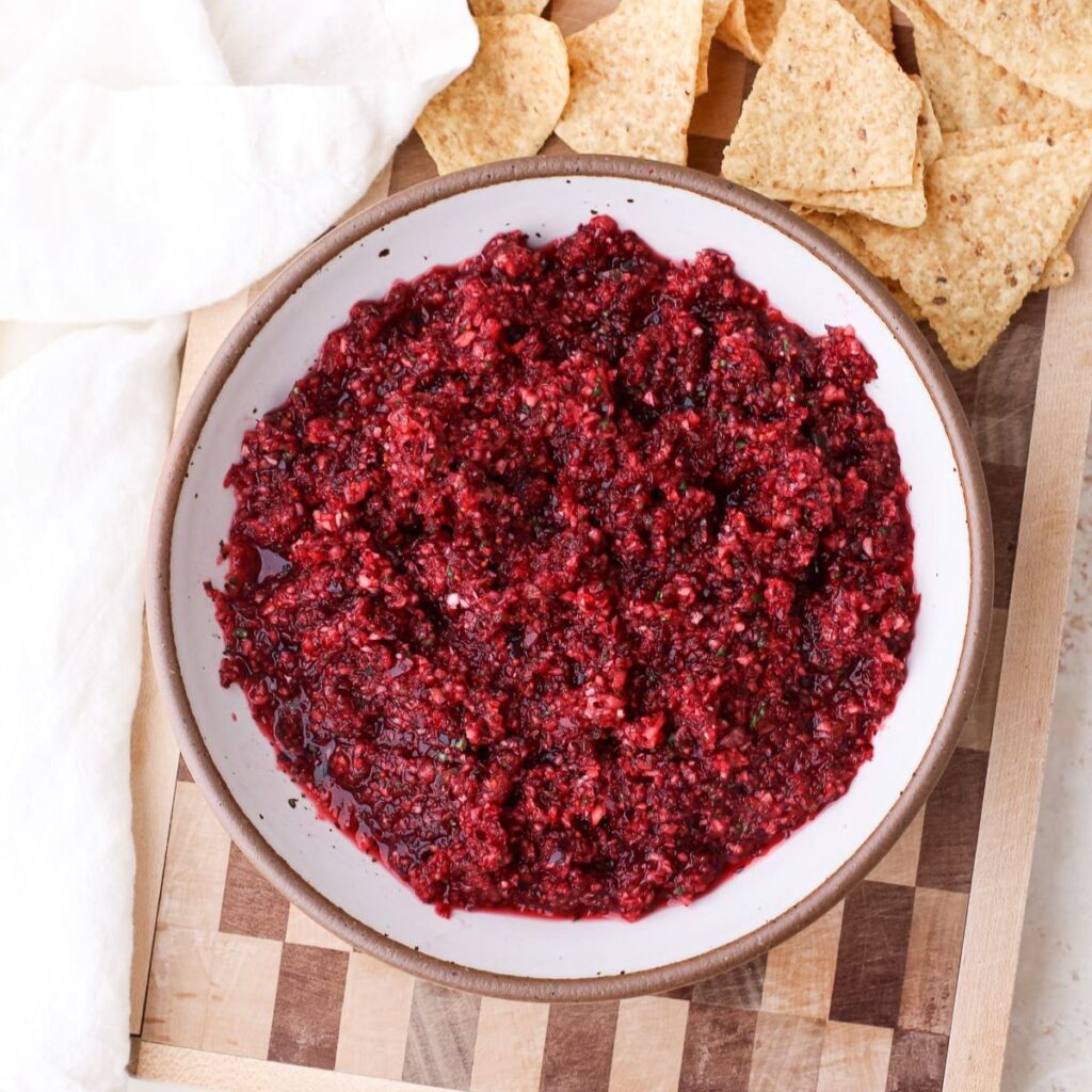 A bowl of fresh cranberry jalapeno salsa on a brown checkered cutting board with tortilla chips.
