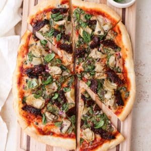 A large homemade mediterranean pizza topped with fresh basil on a striped wooden cutting board cut into 8 pizza slices.