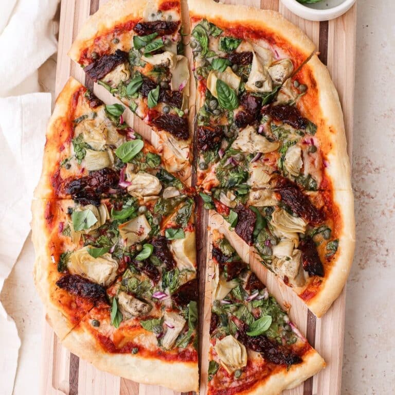 A large homemade mediterranean pizza topped with fresh basil on a striped wooden cutting board cut into 8 pizza slices.