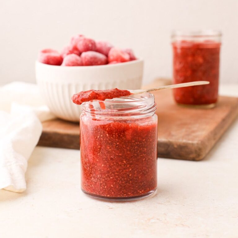 Two jars of homemade strawberry chia jam next to a bowl of frozen strawberries.