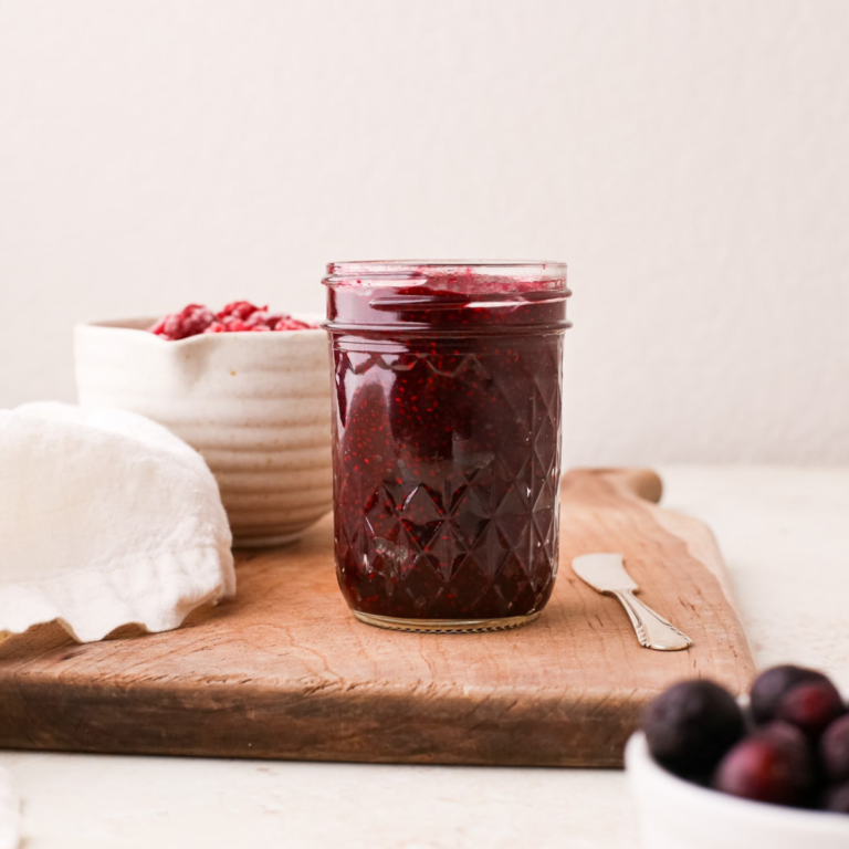 A jar of blue raspberry chia seed jam on a wooden cutting board next to a bowl of raspberries and a bowl of blueberries.