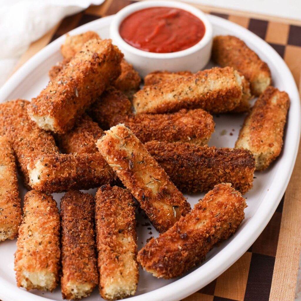 A white oval plate with dairy free mozzarella sticks and a small bowl of red sauce for dipping.