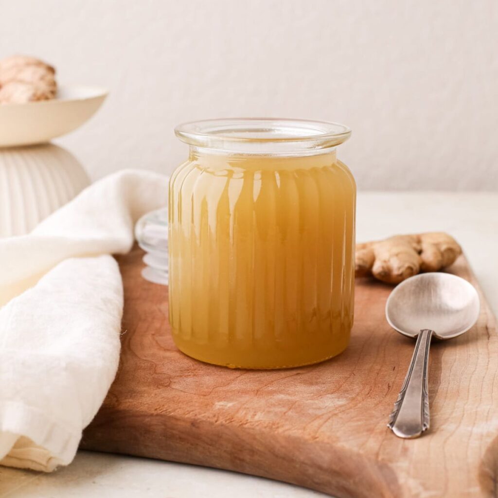 A jar of homemade ginger simple syrup on a cutting board next to fresh ginger root and a spoon.