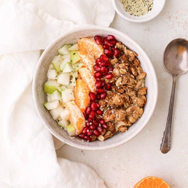 Yogurt bowl topped with granola, pomegranate seeds, orange segments, and cubed fresh pear topped with hemp seeds.