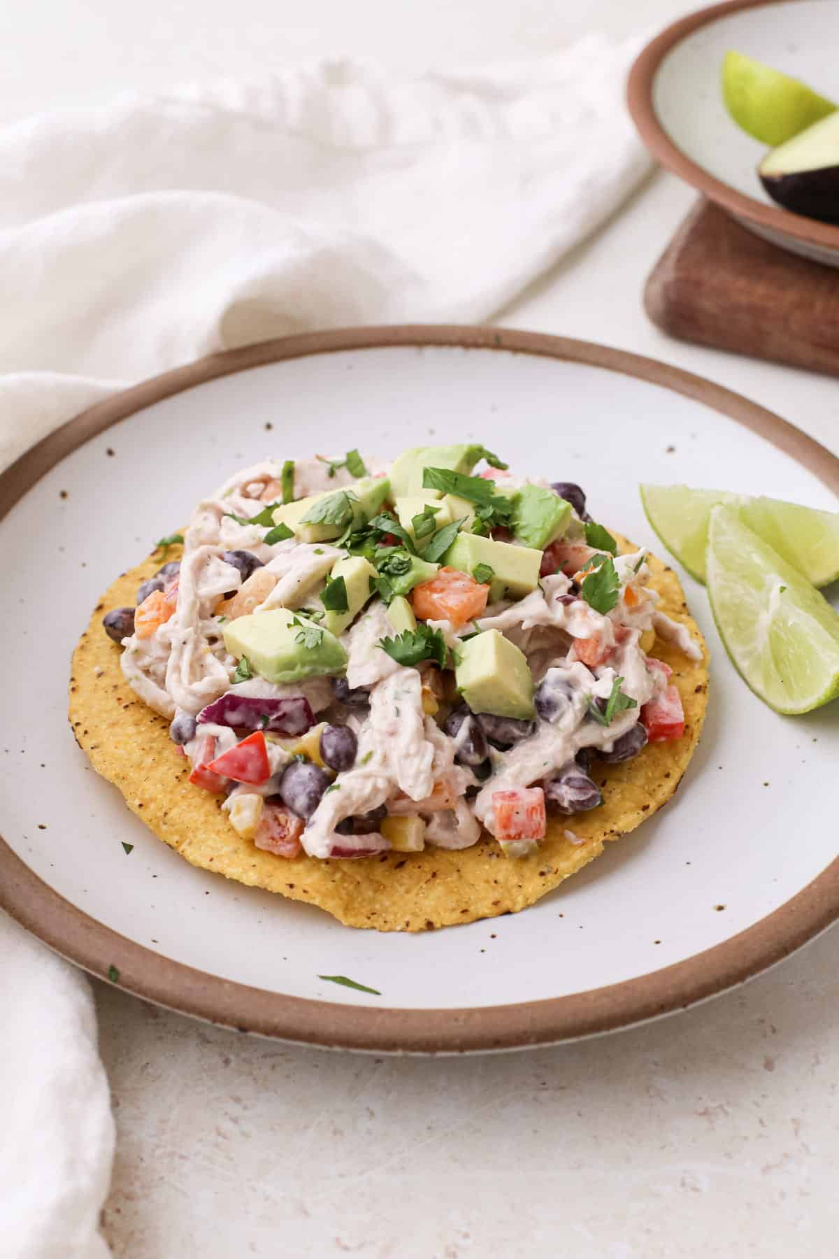 Mexican chicken salad tostadas topped with diced avocado and chopped cilantro on a plate with lime wedges.