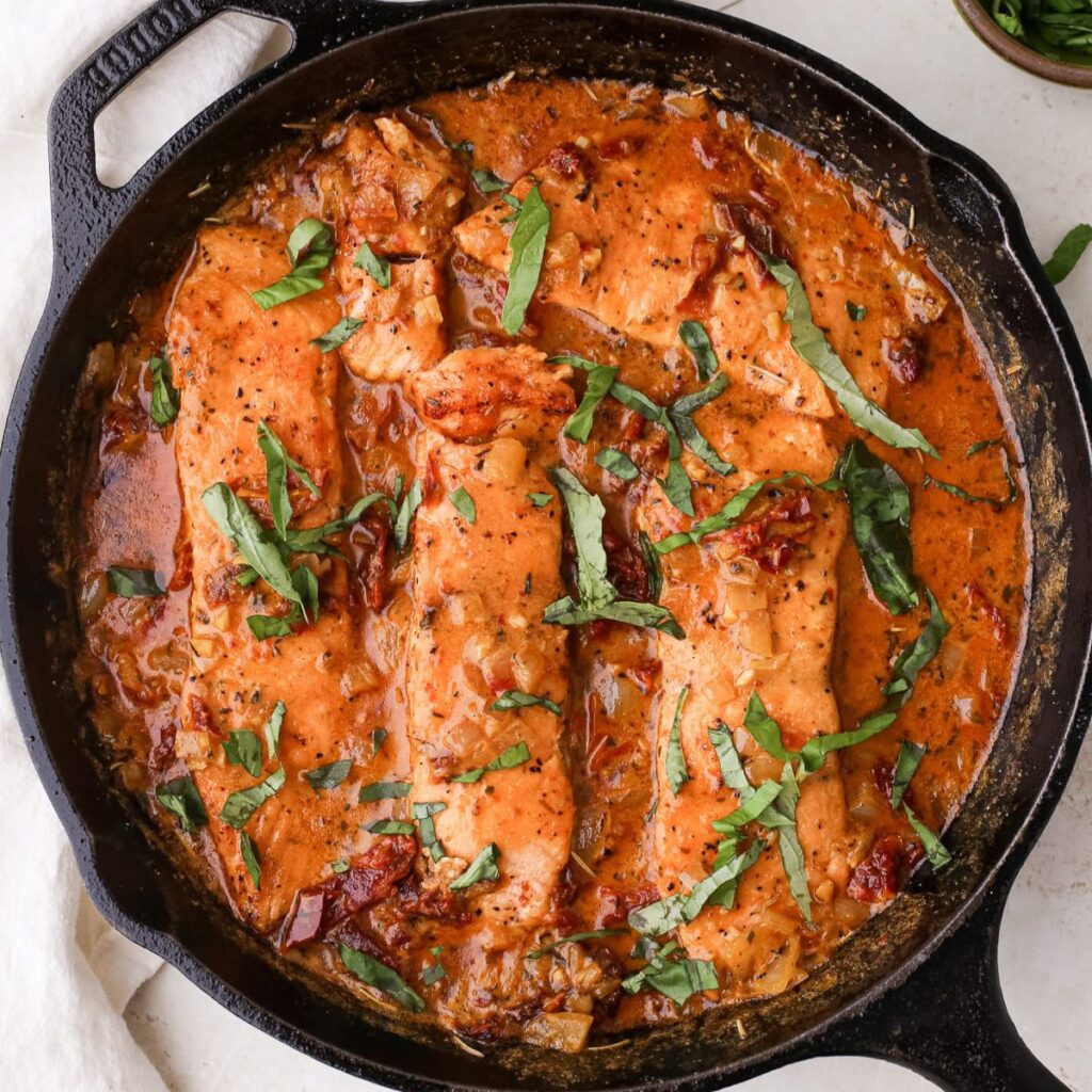 A skillet with marry me salmon in a creamy sun dried tomato sauce topped with chopped fresh basil leaves.
