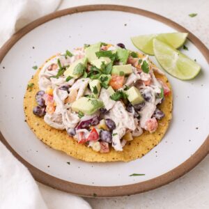 Mexican chicken salad tostadas topped with diced avocado and chopped cilantro on a plate with lime wedges.