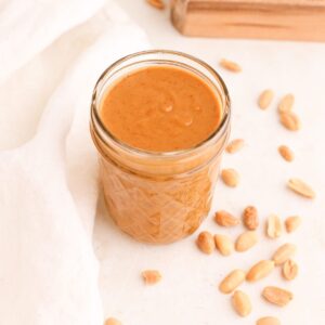 A jar of homemade thai peanut sauce surrounded by roasted peanuts.