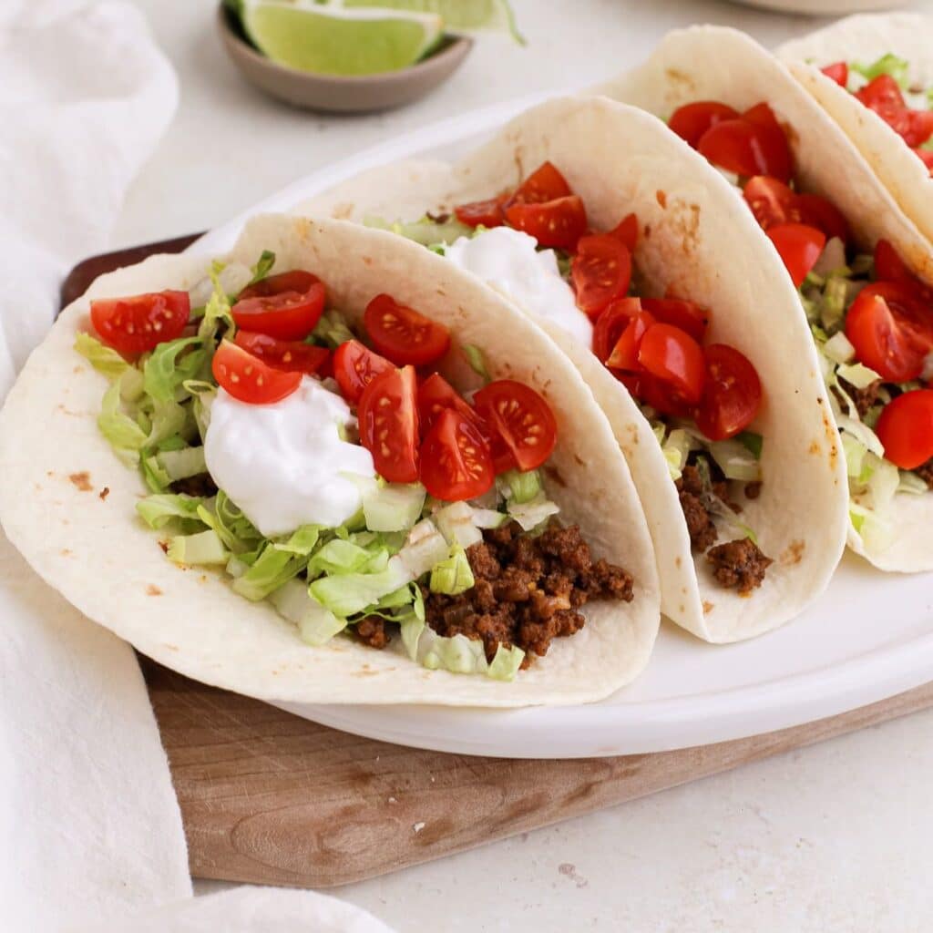 Beef soft tacos on an oval plate topped with shredded lettuce, halved cherry tomatoes, and a dollop of plain yogurt next to a ramekin of fresh lime wedges and more chopped tomatoes.