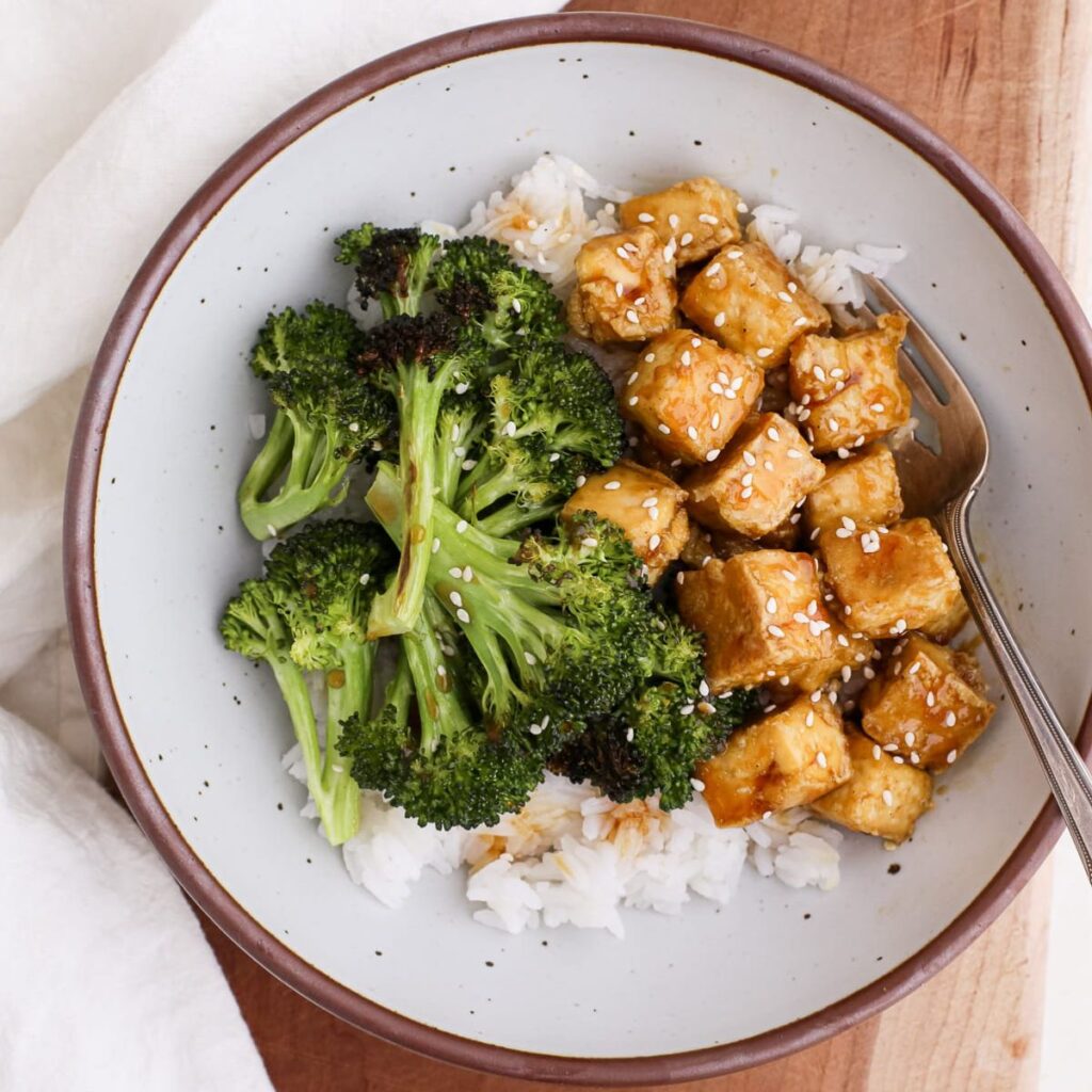 A bowl of honey garlic tofu cubes with roasted broccoli on a bed of white rice topped with sesame seeds.