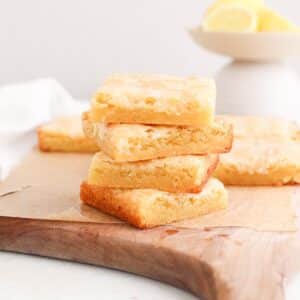 A stack of lemon blondie squares on a strip of parchment paper on a cutting board next to a bowl of fresh lemons.