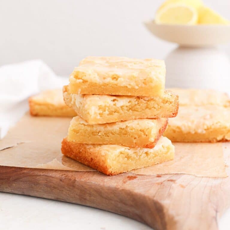 A stack of lemon blondie squares on a strip of parchment paper on a cutting board next to a bowl of fresh lemons.