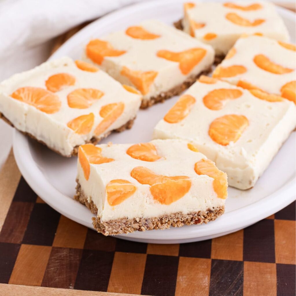 Orange creamsicle frozen yogurt bars with a graham cracker crust topped with mandarin orange segments on an oval plate on top of a checked cutting board.