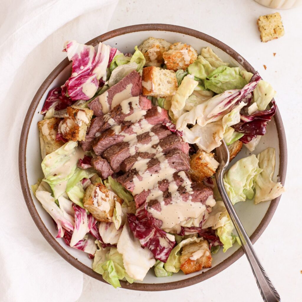 A bowl of caesar salad with sliced steak, homemade croutons, dairy free caesar dressing, and a fork.