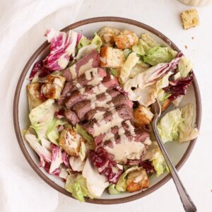 A bowl of caesar salad with sliced steak, homemade croutons, dairy free caesar dressing, and a fork.