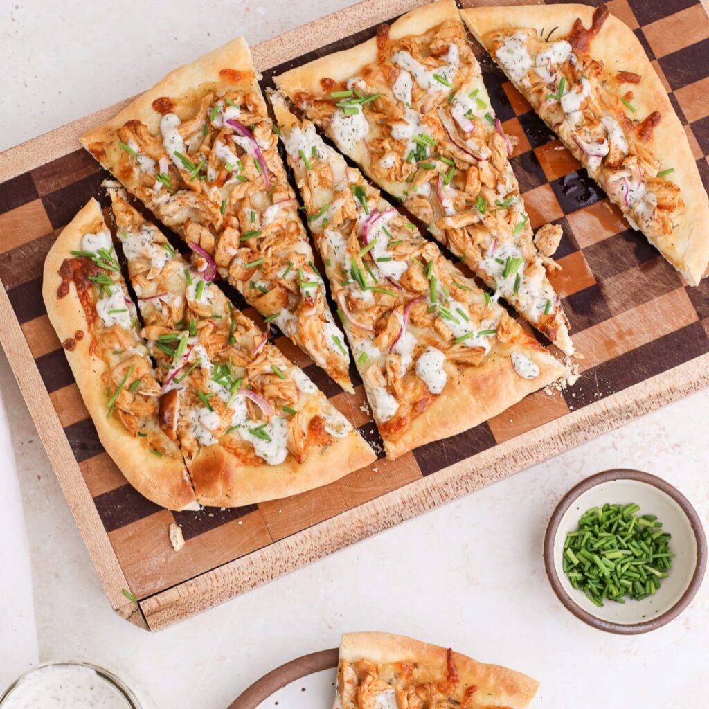 Rectangular buffalo chicken flatbread on a checkered cutting board with one slice on a small plate next to a jar of dairy free ranch dressing a small bowl of chives.