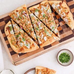 Rectangular buffalo chicken flatbread on a checkered cutting board with one slice on a small plate next to a jar of dairy free ranch dressing a small bowl of chives.