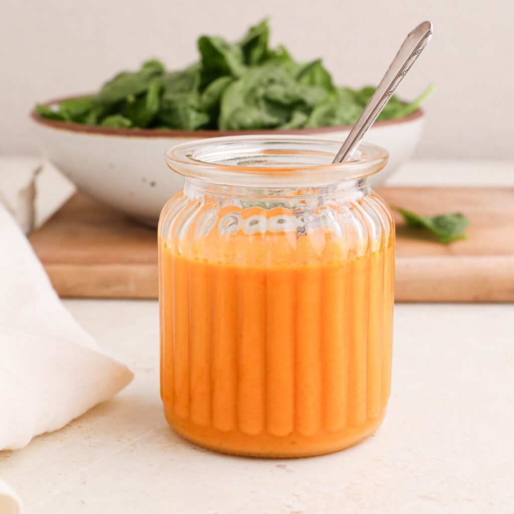 A jar of homemade red curry vinaigrette dressing with a spoon and a bowl of greens on a cutting board.