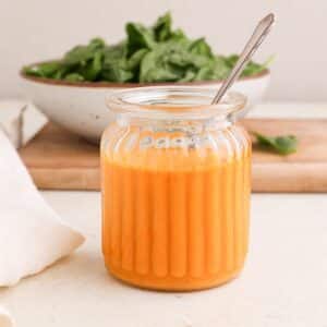 A jar of homemade red curry vinaigrette dressing with a spoon and a bowl of greens on a cutting board.