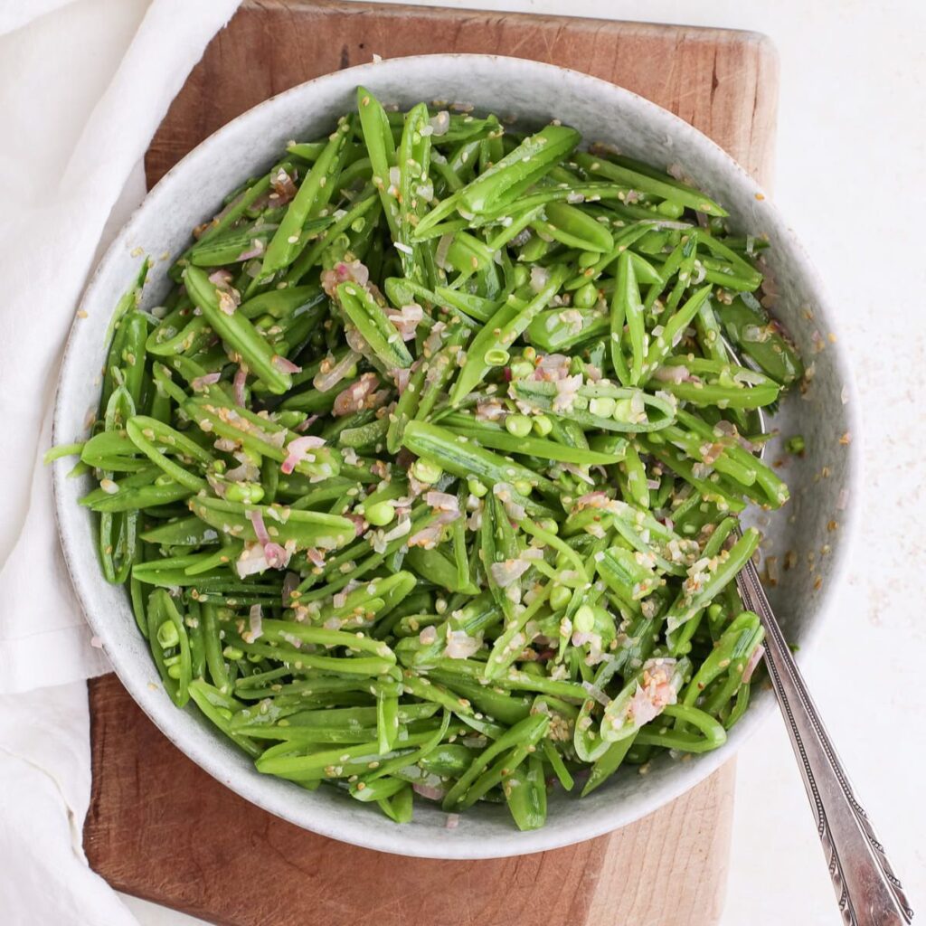 A round serving bowl of snap pea salad with sesame shallot vinaigrette with a spoon in it on a cutting board next to a cloth napkin.