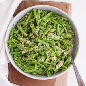 A round serving bowl of snap pea salad with sesame shallot vinaigrette with a spoon in it on a cutting board next to a cloth napkin.