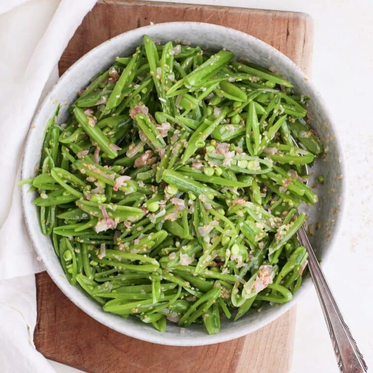 A round serving bowl of snap pea salad with sesame shallot vinaigrette with a spoon in it on a cutting board next to a cloth napkin.