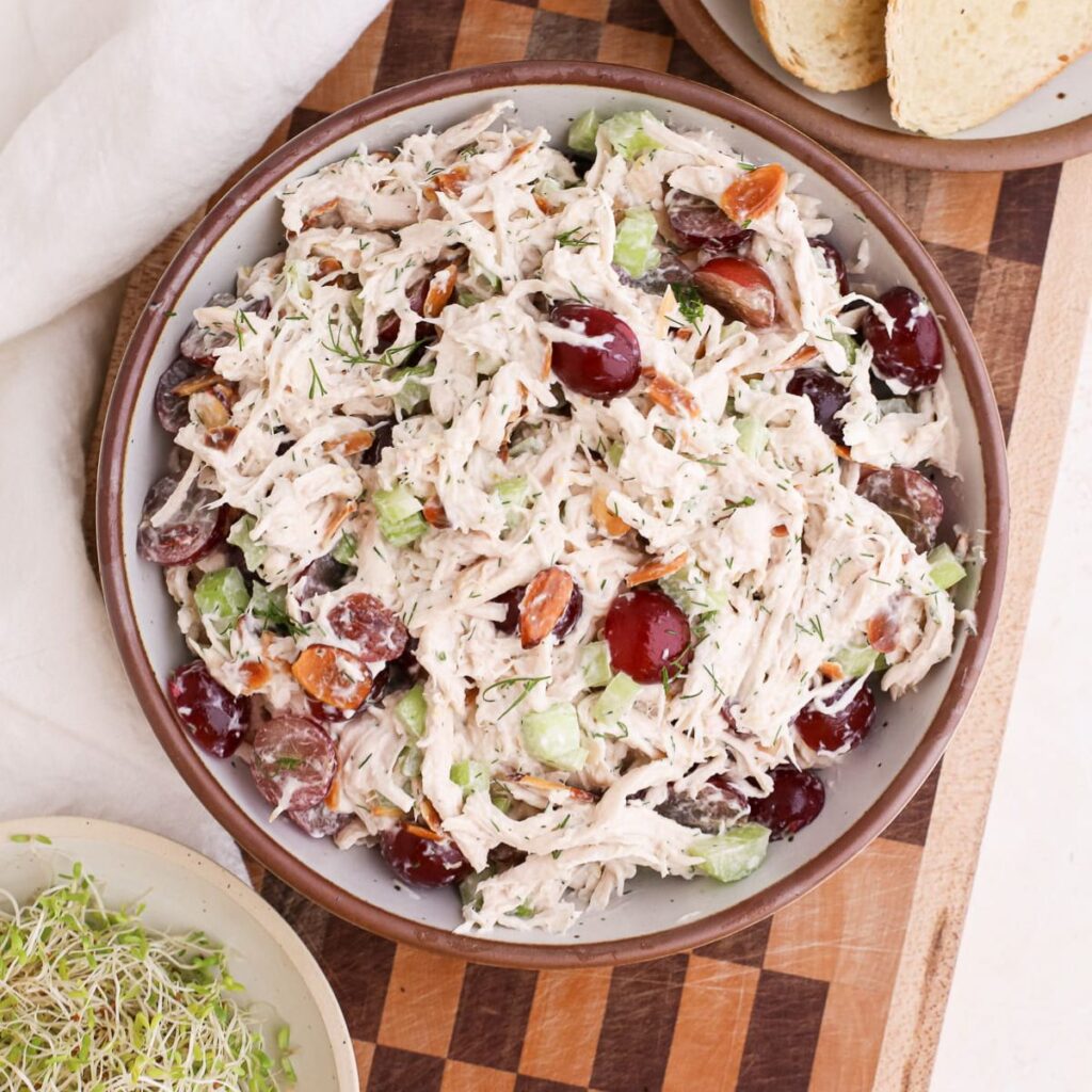 A bowl of homemade chicken salad with red grapes and fresh dill on a checkered cutting board next to toast and sprouts.