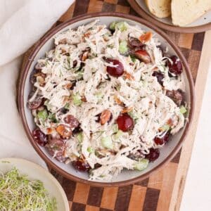 A bowl of homemade chicken salad with red grapes and fresh dill on a checkered cutting board next to toast and sprouts.